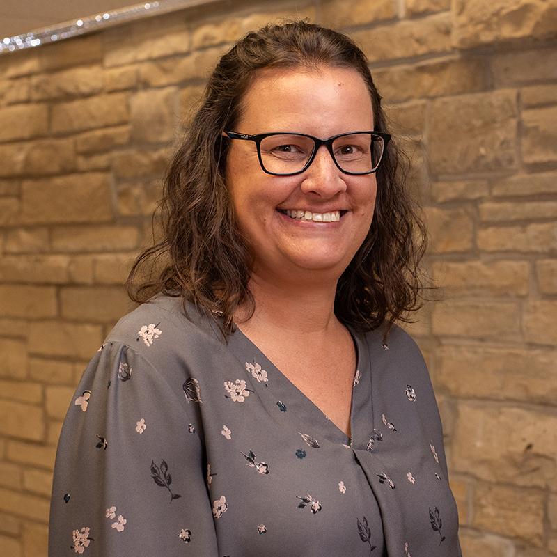 A smiling woman with glasses, wearing a floral blouse, standing against a stone wall backdrop.